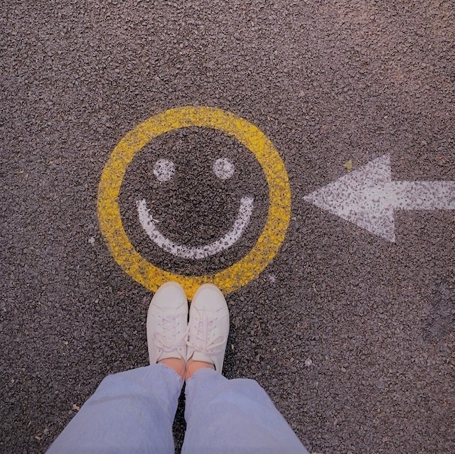Das Bild zeigt einen auf der Straße aufgemalten Smiley, vor dem eine Person steht, von der nur die Füße zu sehen sind