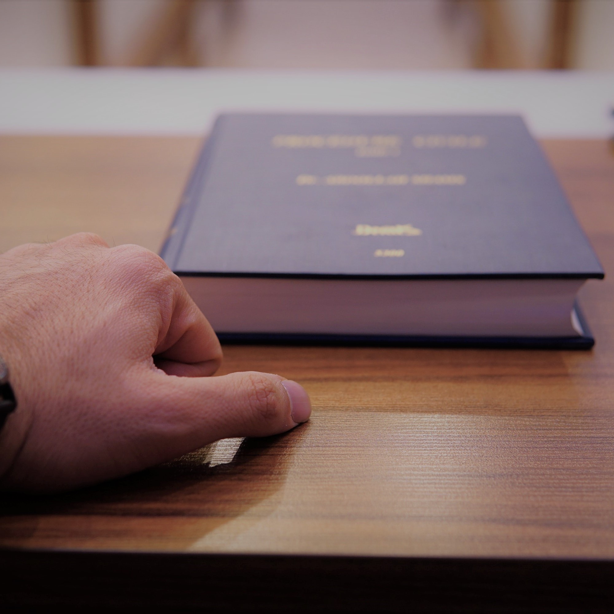Das Bild zeigt die Hand einer männlichen Person auf einem Holztisch. Davor liegt ein Buch.