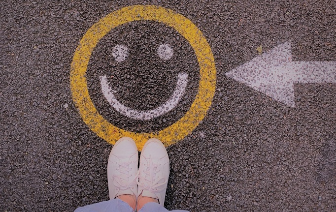 Das Bild zeigt einen Smiley, der auf die Straße gemalt ist und vor dem eine Person steht. Von der Person sind nur die Füße zu sehen.