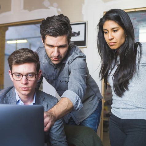 Die Aufnahme zeigt drei Arbeitskollegen im fachlichen Austausch, die in einem Büro gemeinsam einen Laptop betrachten