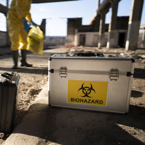 Die Aufnahme zeigt im Vordergrund einen allein stehenden Koffer mit einem Symbol darauf für "Biogefährdung", während im Hintergrund ein Mann mit gelbem Schutzanzug steht 