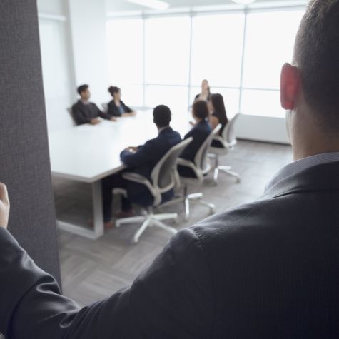 Die Aufnahme zeigt einen Mann, der an der Tür eines Konferenzraumes ein Gespräch von Kolleginne und Kollegen belauscht