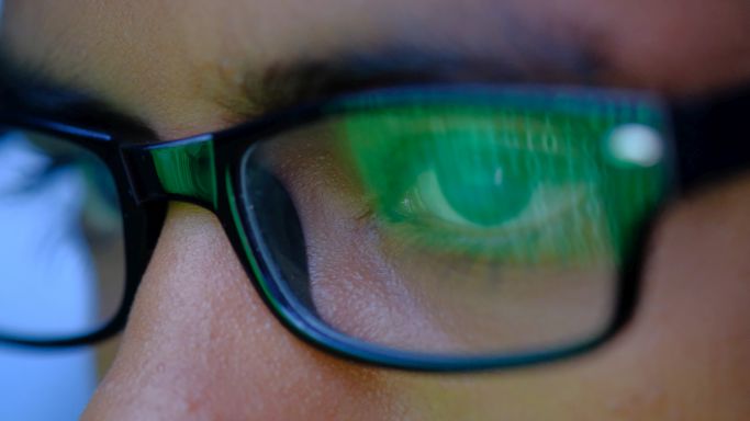 Das Bild zeigt den Ausschnitt eines männlichen Gesichts mit Brille, in dessen Gläsern sich ein binärer Code reflektiert